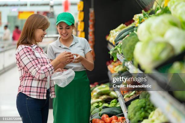 schöne junge mitarbeiterin hilft weiblichen kunden bei der auswahl von gemüse im supermarkt - supermarket help stock-fotos und bilder