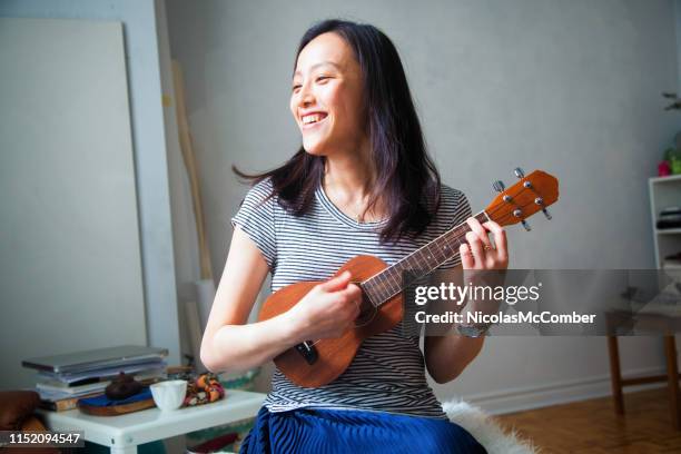 jeune femme asiatique de millénaire ayant l’amusement jouant le ukulélé dans son appartement de studio - pincer la corde photos et images de collection