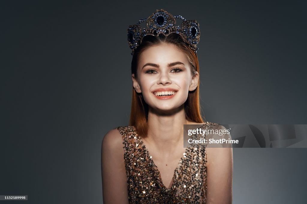 The woman in the glittering dress and the crown is smiling beautifully