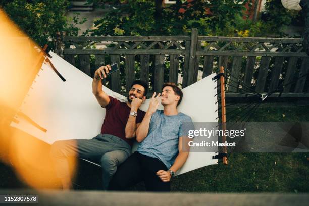 high angle view of smiling friends taking selfie on smart phone while resting in hammock at yard - cell mates stock-fotos und bilder