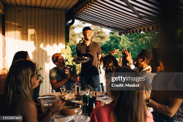 friends at dining table clapping for man serving food in dinner party - sonnensegel stock-fotos und bilder