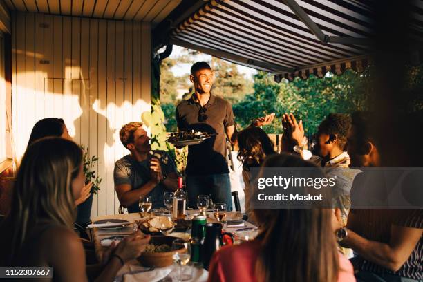 friends at dining table clapping for man serving food in dinner party - barbecue stock-fotos und bilder