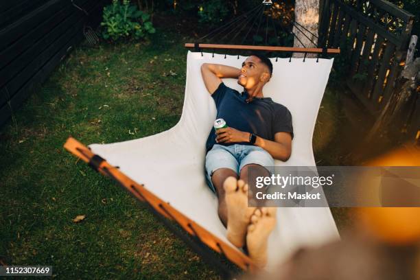 high angle view of young man having drink while lying in hammock at back yard - trädgård hängmatta bildbanksfoton och bilder