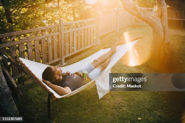 high angle view of young man with drink lying on hammock in back yard - hammock stock pictures, royalty-free photos & images