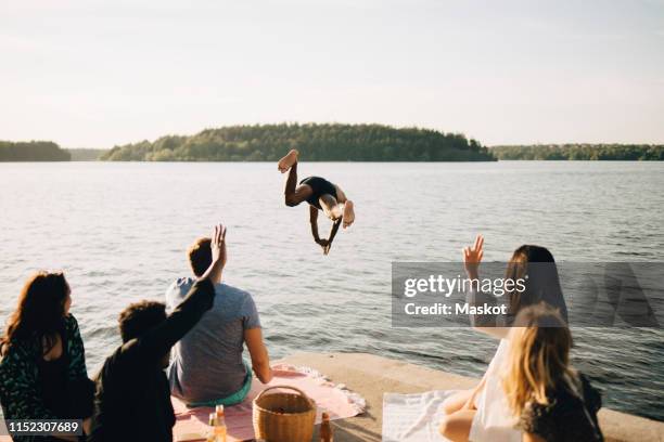 men and women looking at shirtless friend diving from jetty at lake against sky - stockholm summer stock-fotos und bilder