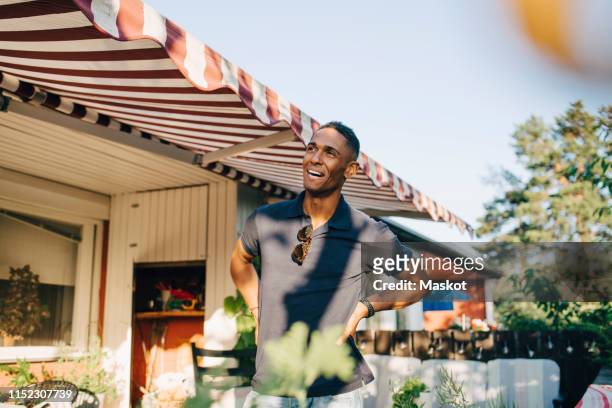 happy young man looking away while standing with hands on hip at yard - striped awning stock pictures, royalty-free photos & images
