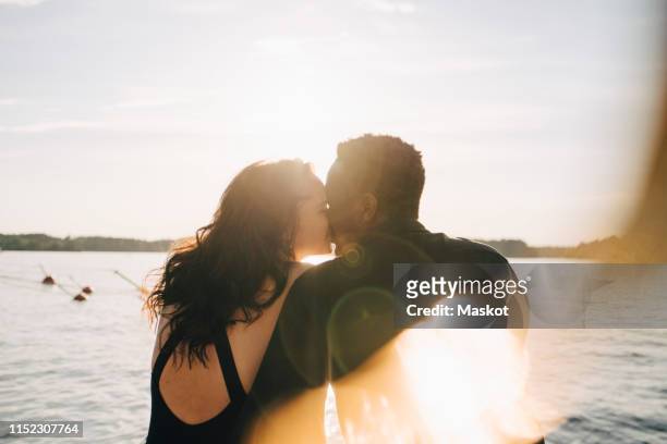 multi-ethnic couple kissing while sitting against lake during summer - romance photos et images de collection