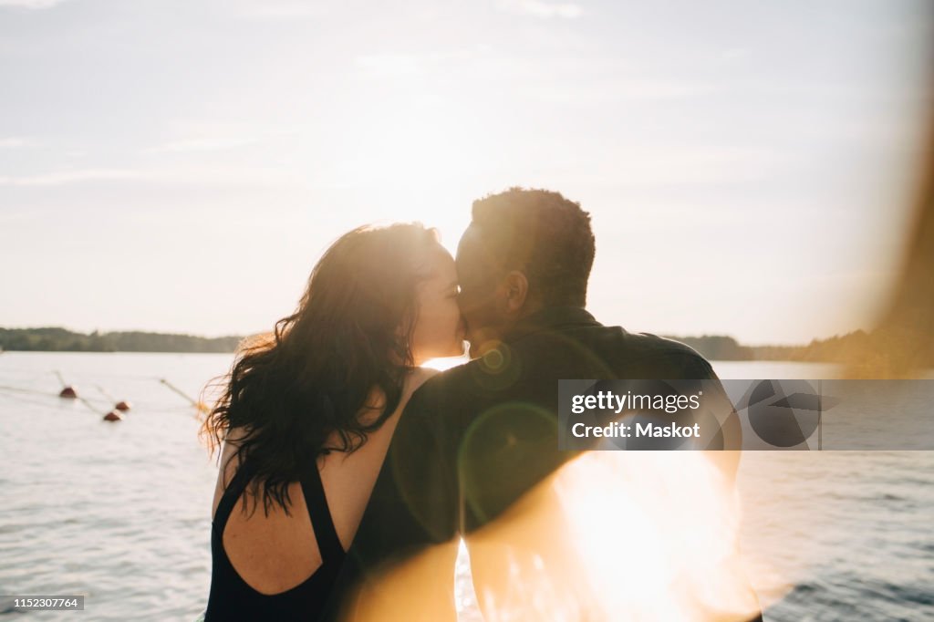 Multi-ethnic couple kissing while sitting against lake during summer