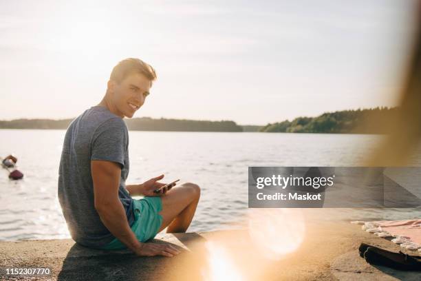 portrait of smiling young man sitting with mobile phone on jetty by lake - scandinavia picnic stock pictures, royalty-free photos & images