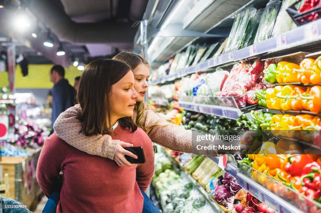 Cute daughter choosing bell peppers while being piggybacked by mother in store