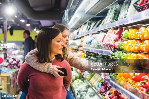 cute daughter choosing bell peppers while being piggybacked by mother in store - food shopping stock pictures, royalty-free photos & images
