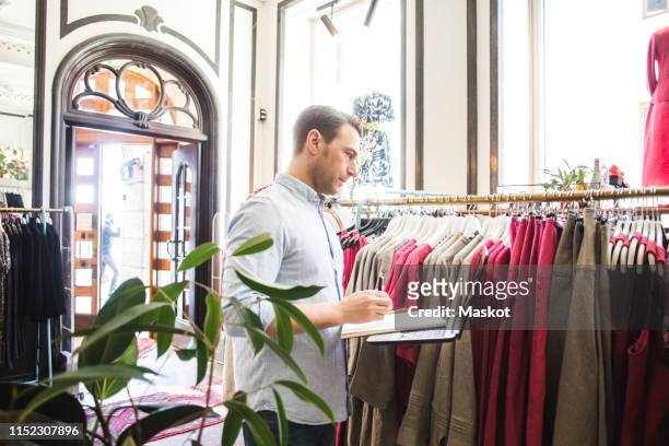 side view of sales clerk looking at clothes rack while holding book and laptop in boutique - clothes hanging on rack at store for sale foto e immagini stock