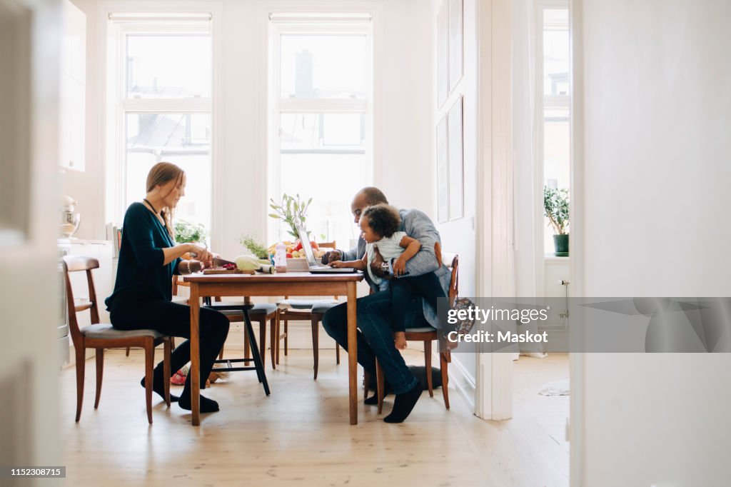 Father showing laptop to daughter while mother cutting vegetables at table in house