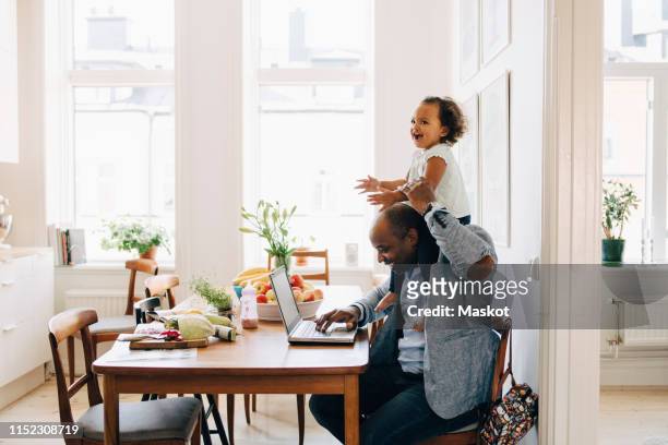 father carrying cheerful daughter on shoulder while working on laptop at table in house - carrying on shoulders stock pictures, royalty-free photos & images