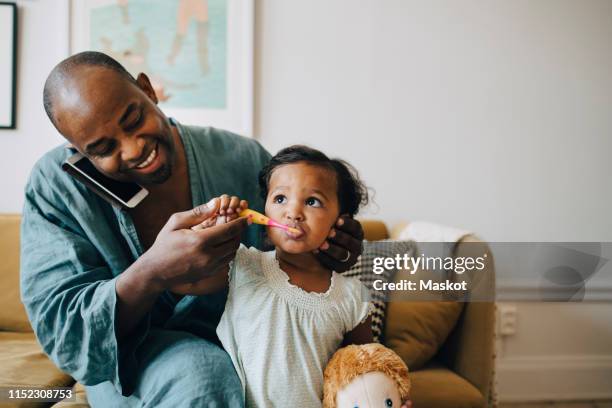 man talking on mobile phone while brushing daughter's teeth at home - brush teeth phone stock-fotos und bilder