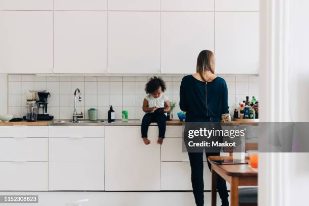 full length of girl using smart phone while mother working in kitchen at home - femme de dos smartphone photos et images de collection