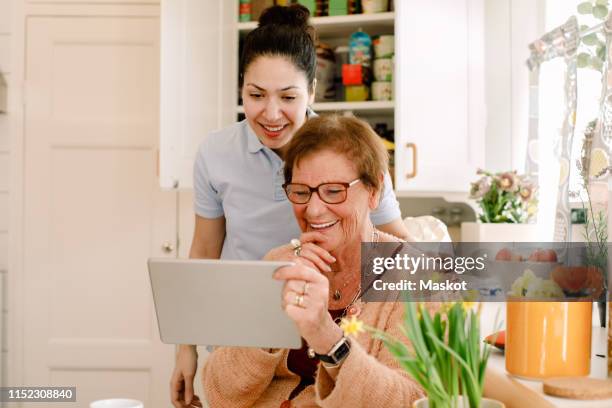 smiling elderly woman and young female caregiver looking at digital tablet in nursing home kitchen - carer allowance stock pictures, royalty-free photos & images