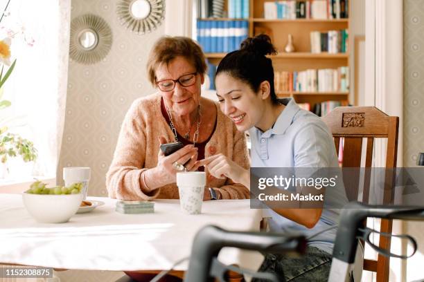 smiling young female social service worker assisting retired elderly woman using smart phone at table in nursing home - volunteer aged care stock pictures, royalty-free photos & images