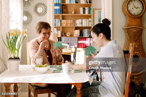 smiling young female volunteer playing cards with retired elderly woman at table in nursing home - retirement card stock pictures, royalty-free photos & images