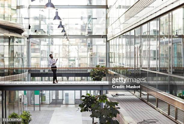 full length of young businessman standing at atrium in office - businessman distance window ストックフォトと画像