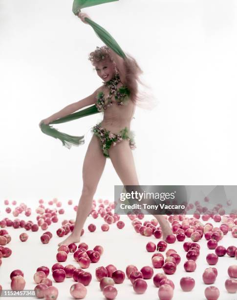 Dancer and choreographer Gwen Verdon is dancing wearing green bikini holding a green scarf on a floor covered with red apples.