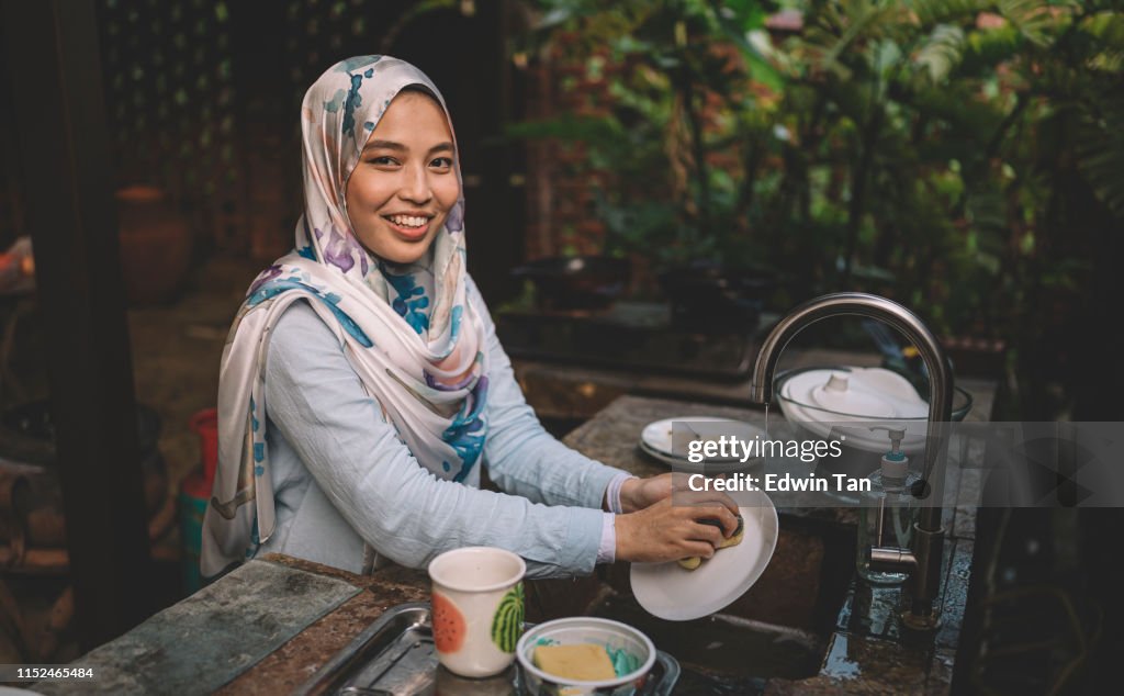 A Malay lady cleaning and washing dishes after dinner party looking at camera