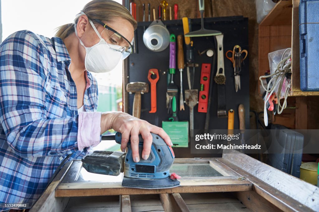Rijpe vrouw Upcycling meubilair in workshop thuis met behulp van elektrische schuurmachine