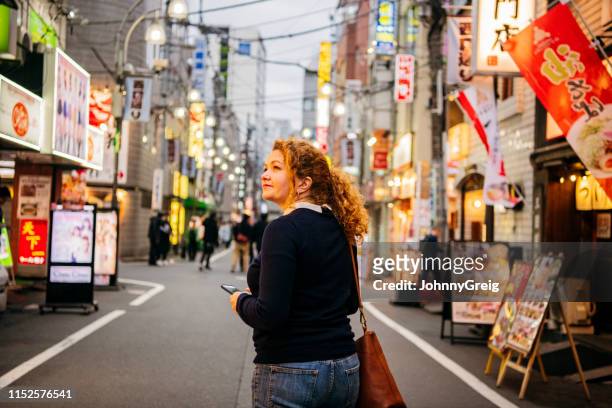 woman walking along tokyo street holding mobile phone - visitor attractions stock pictures, royalty-free photos & images