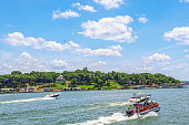 Pontoon boat full of people and two speedboats race down lake with luxury homes and docks on shore under bright blue sky with clouds and helicopter above