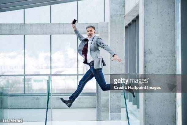 a mature businessman with smartphone in a modern office, jumping. copy space. - gray jacket stock pictures, royalty-free photos & images
