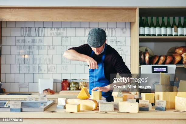 cheesemonger cutting cheese with cheese wire surrounded by a variety of cheese on counter top - デリカッセン ストックフォトと画像