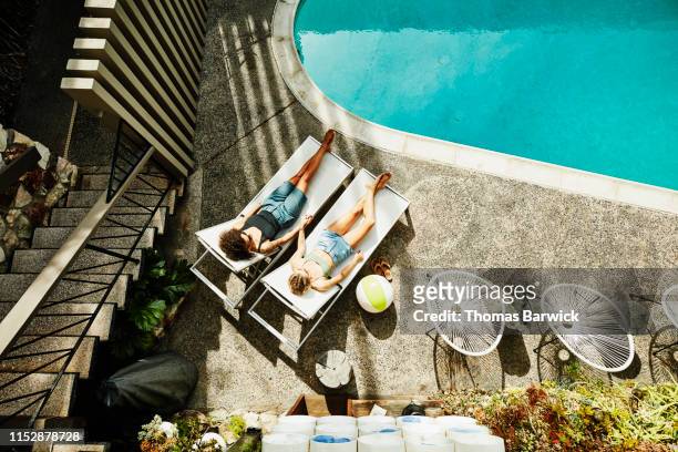 overhead view of lesbian couple holding hands while relaxing in lounge chairs by hotel pool - ideal wife stock pictures, royalty-free photos & images