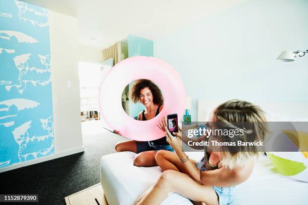 woman taking photo with smart phone of smiling wife holding up pool toy while relaxing on bed in hotel room - funny lesbian 個照片及圖片檔