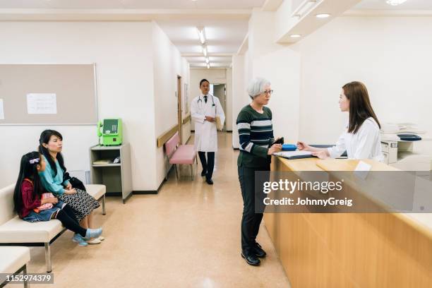 senior woman talking to receptionist in hospital waiting area - hospital reception stock pictures, royalty-free photos & images