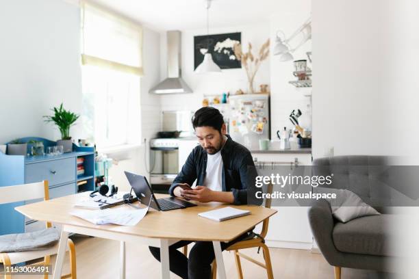 hombre asiático usando teléfono inteligente mientras trabaja desde casa - autonomo smartphone tablet fotografías e imágenes de stock