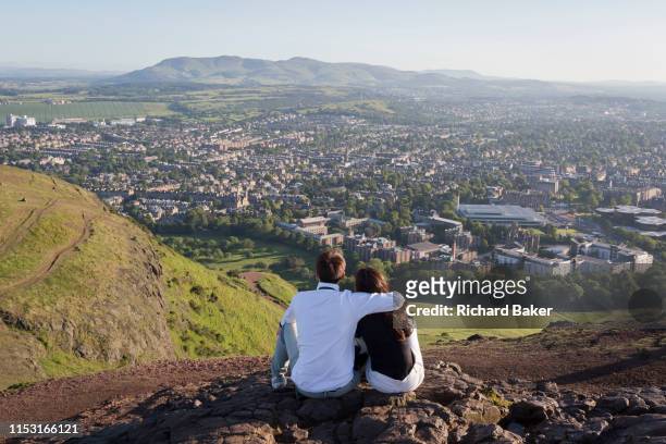 Young couple gaze out towards the city of Edinburgh from the summit of Arthur's Seat in Holyrood Park, on 26th June 2019, in Edinburgh, Scotland....