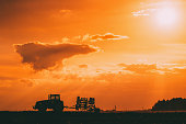 Tractor Rides On Countryside Road. Beginning Of Agricultural Spring Season. Cultivator Pulled By A Tractor In Rural Field Landscape Under Sunny Summer Sunset Sunrise Sky. Backlit Dramatic Lighting