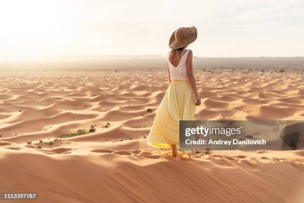 a young caucasian woman in a long skirt, shirt and straw hat standing on top of a sand dune and looks towards the sunrise. sunrise in the merzouga (sahara) desert. - morocco stock pictures, royalty-free photos & images