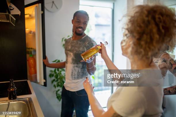 young man opening fridge and passing the beer to his flatmate - beverage fridge stock pictures, royalty-free photos & images