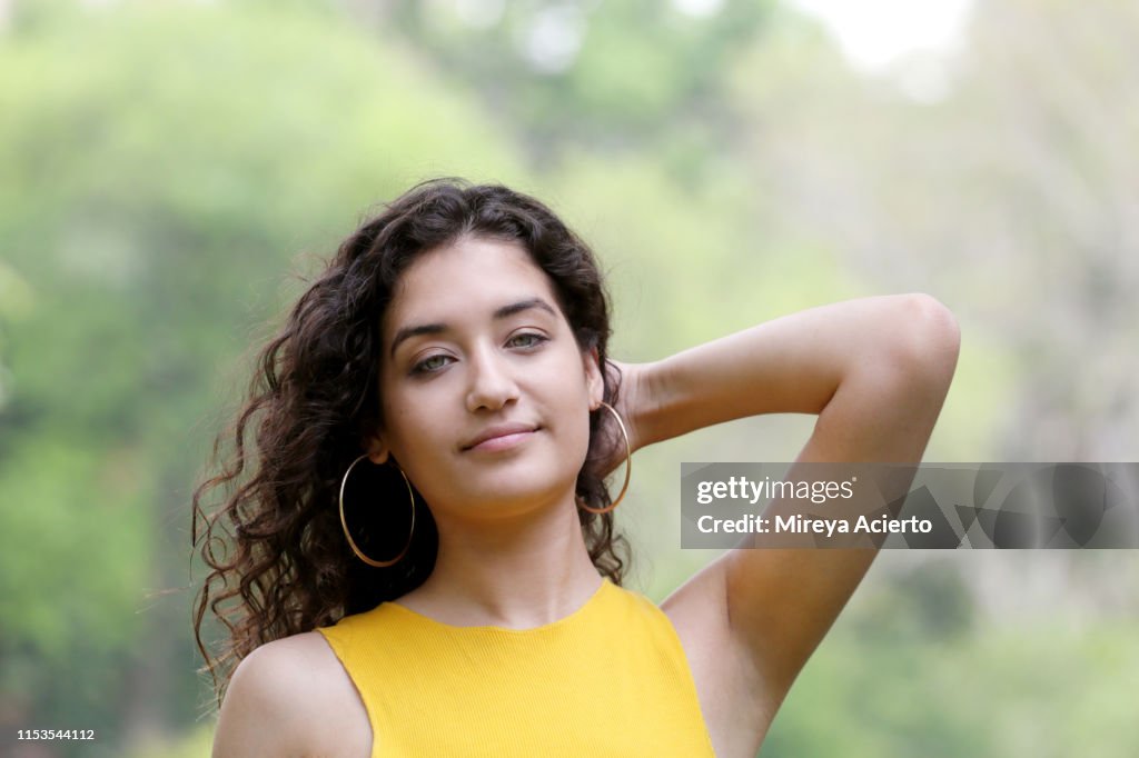 A portrait of a LatinX female millennial wearing a yellow top, stands in a public park.