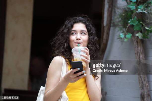 a latinx female millennial smiles while holding a cell phone and drinking an iced coffee, on a summer day in new york city. - ice coffee drink stock pictures, royalty-free photos & images