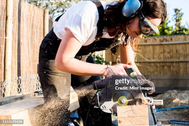 woman wearing protective goggles and ear protectors holding circular saw, cutting piece of wood on building side. - elektrisch gereedschap stockfoto's en -beelden