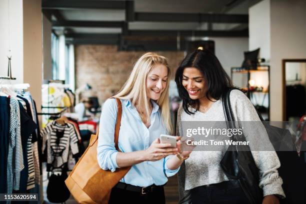 two womem looking at smartphone while clothes shopping - frauen shoppen stock-fotos und bilder