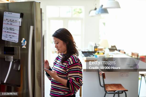 mid adult woman checking food in fridge holding tablet - asian woman shopping grocery stock pictures, royalty-free photos & images
