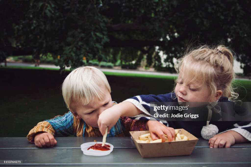 Two toddlers sharing hot chips and tomato sauce at a picnic in the park