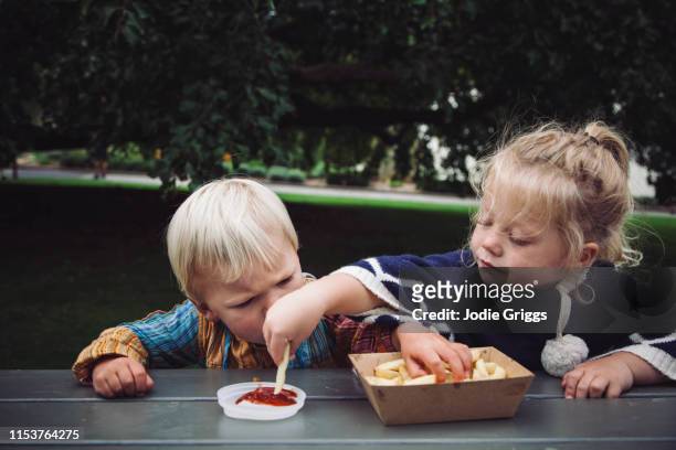 two toddlers sharing hot chips and tomato sauce at a picnic in the park - kind stock pictures, royalty-free photos & images