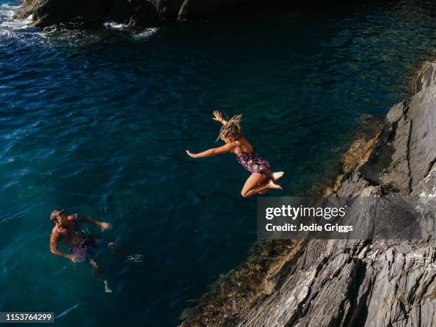 father watching young girl jumping off a rock into the ocean - natural shot female stock-fotos und bilder