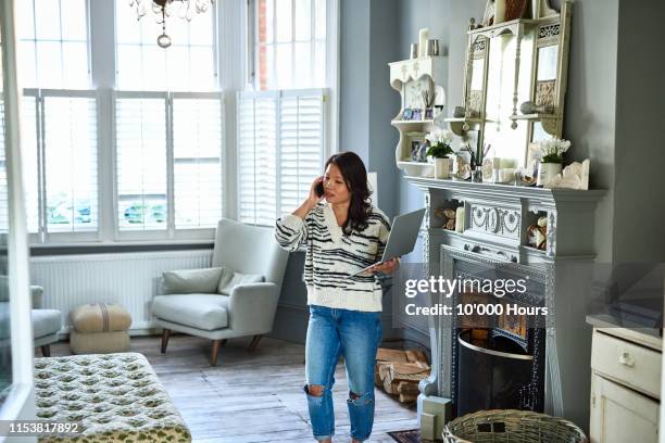 mid adult woman on phone with laptop in living room - patience office stock pictures, royalty-free photos & images