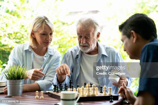 senior man playing chess and thinking about the game with her woman help with young man . he 's preparing to move chess figurine. - 老人痴呆症 個照片及圖片檔