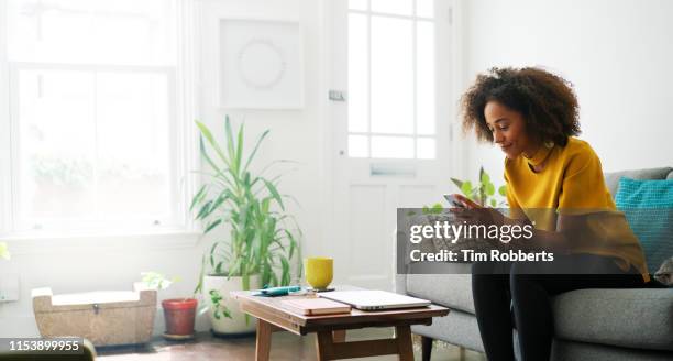 woman sat on sofa using smart phone - woman using smartphone with laptop stock-fotos und bilder